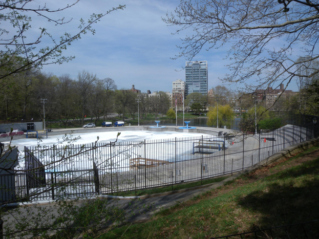 Eisstadion in einem Park umgeben von einem Metallzaun, Gras, Bäumen, Fahrzeugen, Straßenlaternen, einem Gewässer, Gebäuden und einem bewölkten Himmel.