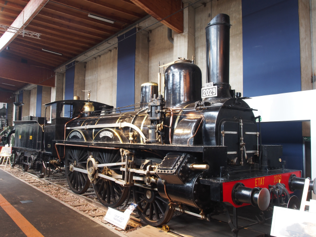 Eine große, alte Dampflokomotive, die auf Eisenbahnschienen in einem Museum ausgestellt ist, umgeben von Informationsschildern, Ständen und anderen Ausstellungen, mit einer Wand und Deckenbeleuchtung im Hintergrund.