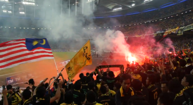 Eine große Menge in einem Stadion mit Fackeln, Fahnen und einer Kamera, mit Rauch und Schildern, die "Malaysia vs Malaysia Live Streaming" im Hintergrund zeigen.