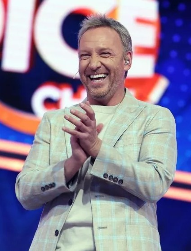 Ein Mann in einem Anzug, der auf einer Bühne klatscht, mit dem 'The Voice'-Schild im Hintergrund.