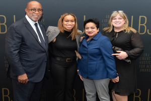 Vier Personen auf einem roten Teppich, die lächeln und für ein Foto posieren, mit einem Schild im Hintergrund, auf dem 'Liberty Justice for All Awards' steht.