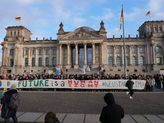 Eine Gruppe von Menschen steht vor dem Reichstaggebäude in Berlin und hält ein Transparent mit der Aufschrift "Zukunft ist ein Human Neustar ima" und ist von Flaggen umgeben.