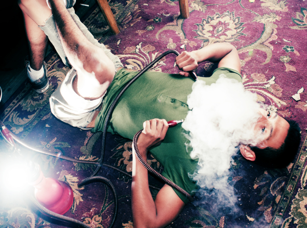 Ein Junge in einem grünen T-Shirt liegt auf einem roten Teppich und raucht einen Hookah, mit einem roten Hookah-Rohr und Rauch daneben.