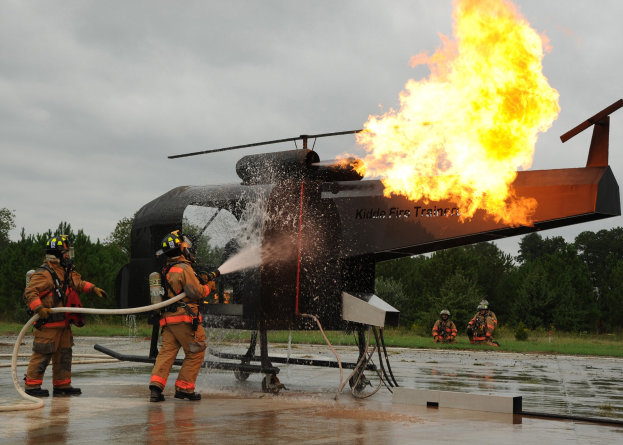 Feuerwehrleute mit Helmen spritzen Wasser auf einen brennenden Hubschrauber, während im Hintergrund Trees und ein klarer blauer Himmel zu sehen sind.
