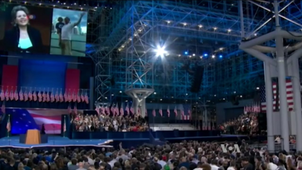 Große Menschenmenge vor einer Bühne mit einem Rednerpult, Mikrofon, Flaggen, einem Overhead-Bildschirm und Beleuchtungskörpern bei dem Anschein eines Präsidentschaftsdebattens.