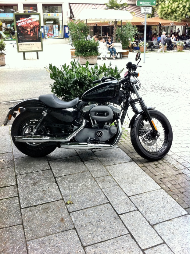 Ein schwarzes Fahrrad steht auf einem Boden mit einer Pflanze dahinter und einem Plakat weiter hinten; rechts davon ein Geschäft mit Dach, vor dem Zelte stehen, darunter Menschen sitzen und ein Schild an einem Pfahl; ein Baum ist oben rechts und ein Gebäude mit Glastüren dahinter links vom Plakat.