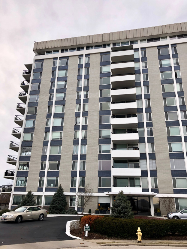 Hohes Wohnhochhaus mit zahlreichen Fenstern, einem roten Feuerhydranten im Vordergrund, Fahrzeugen auf der Straße, Grünfläche einschließlich Pflanzen und Bäumen sowie einem bewölkten Himmel im Hintergrund.