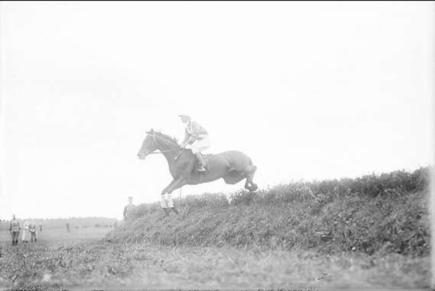 Schwarzes und weißes Foto eines Pferdes und Reiters, die über einen grasbewachsenen Hügel springen, während das Pferd galoppiert und der Reiter einen Helm und einen Mantel trägt, während Zuschauer im Hintergrund beobachten.