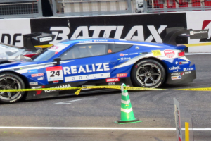 Ein blauer und weißer Rennwagen fährt auf einer Rennstrecke, mit einem Verkehrskegel im Vordergrund und Schildern mit Text, Geländern und anderen Gegenständen im Hintergrund.