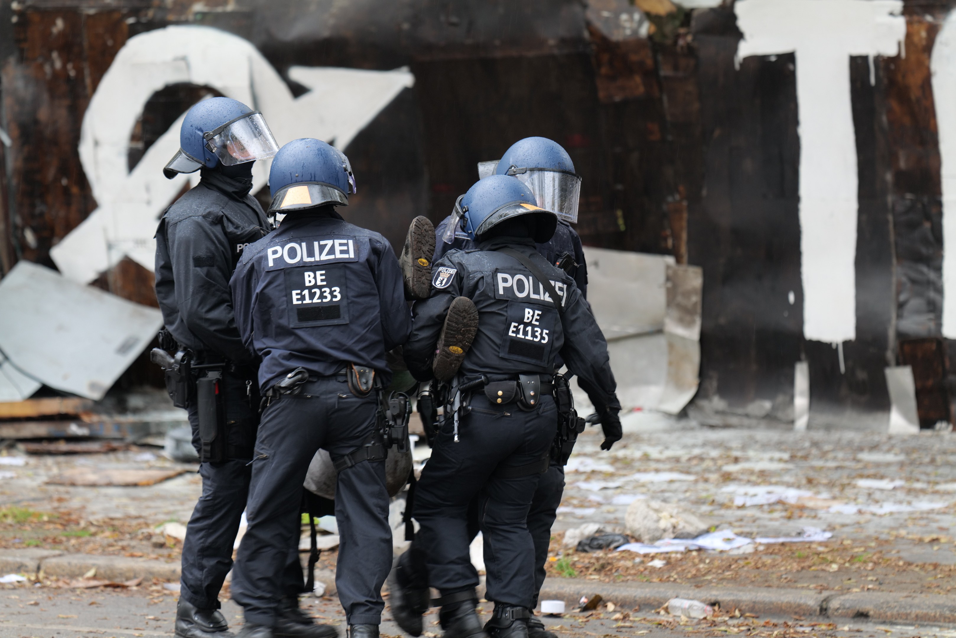 Polizeibeamte in schwarzen Uniformen und blauen Helmen stehen vor einem Gebäude mit Graffiti während einer Ausschreitung, mit verstreuten Papieren und Gegenständen auf dem Boden.