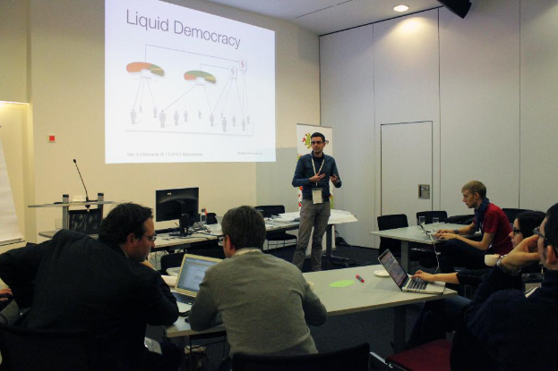 Fünf Personen in einem Konferenzsaal, die einem stehenden Redner zuhören, mit Tischen, auf denen Laptops, Marker, Papiere, ein Monitor, ein Mikrofon und ein Projektor mit Text auf der Wand zu sehen sind.
