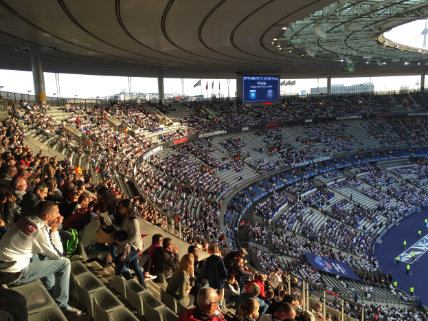 Eine große Menschenmenge sitzt im Allianz Stadion in München, Deutschland, und schaut ein Fußballspiel. Rechts im Bild befindet sich eine Bühne mit ein paar Leuten darauf und im Hintergrund sind Fahnen, Stangen und ein Bildschirm zu sehen. Der Himmel ist oben im Bild zu sehen und das Stadion scheint das Allianz Stadion in München, Deutschland, zu sein.