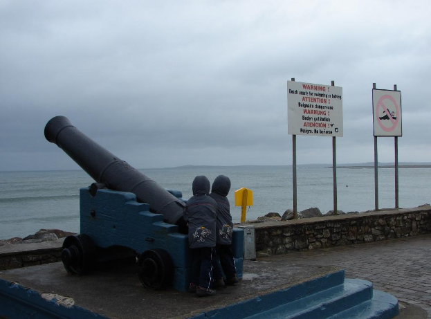 Eine militärische Waffe wird in der Nähe von Kindern, Schildern, einer Felswand und Pfählen gezeigt, mit einem bewölkten Himmel und Wasser im Hintergrund.