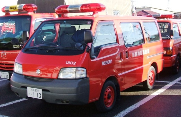 Drei rote Rettungswagen mit weißen und schwarzen Markierungen nebeneinander auf einem Parkplatz geparkt, mit Gebäuden und einem klaren Himmel im Hintergrund.