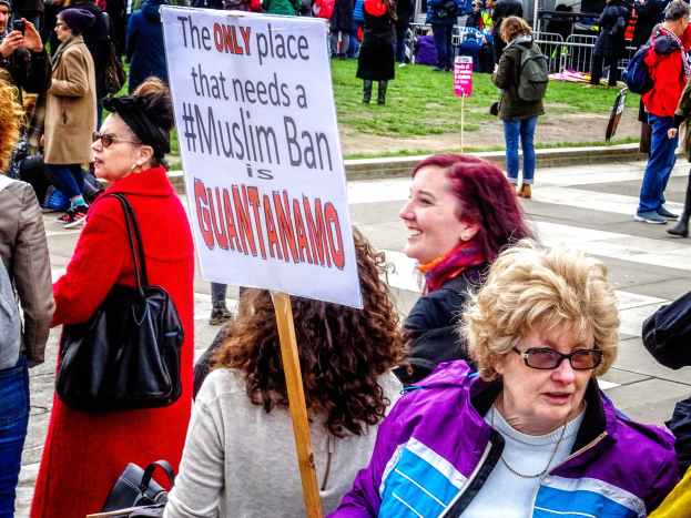 Eine Gruppe von Menschen steht auf dem Boden, einige halten Taschen, eine Frau hält ein Schild mit der Aufschrift "Der einzige Ort, an dem ein Muslimverbot benötigt wird, ist Guantanamo". Im Hintergrund sehen wir einen Metallzaun, Gras, ein Schild mit Text, einige Pfosten, ein Gebäude mit Fenstern und Bäume.