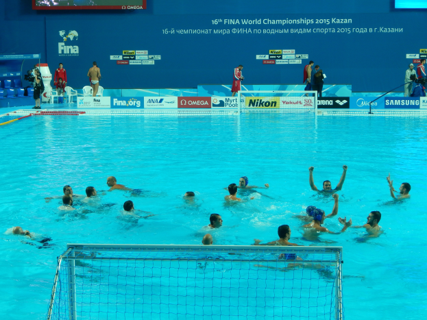 Eine Gruppe von Menschen, die Wasserball in einem Pool mit einem Netz spielen, Zuschauer und Tafeln mit "FINA World Championships 2015 Kazan"-Text im Hintergrund.