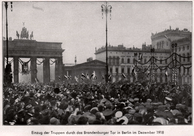 Eine große Menge Menschen mit Hüten und Fahnen steht vor dem Brandenburger Tor in Berlin, Deutschland im Jahr 1918, mit dem Torbogen, Säulen, Statuen und umliegenden Gebäuden im Hintergrund und Text am unteren Bildrand.