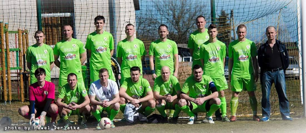 Eine Gruppe von Männer in grünen T-Shirts steht und hockt auf einem Fussballfeld mit einem Ball vor ihnen, ein Torpfosten rechts und ein Hintergrund aus Bäumen, Geb&aumluden, Masten, Fahrzeugen und dem Himmel.