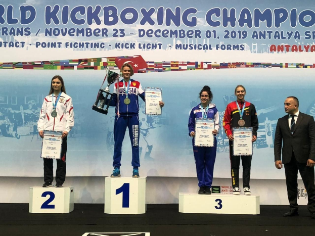Fünf Personen mit Trophäen und Urkunden auf einem Podium mit einem "World Kickboxing Championship Antalya 2019"-Banner im Hintergrund.
