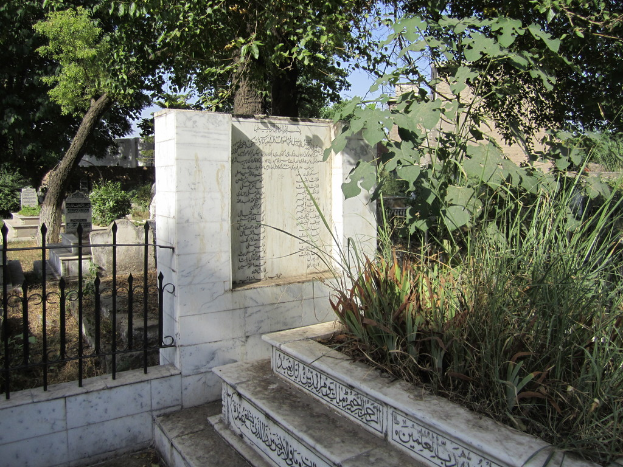 Ein Blick auf einen Friedhof mit Treppe unten, Gras und Bäumen rechts, einer Gedenktafel in der Mitte, einem Geländer daneben, Gräbern dahinter und Bäumen und Gebäuden im Hintergrund.