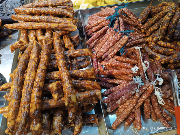 Ein Tisch mit verschiedenen Wurstsorten in Schalen, umgeben von anderen Lebensmitteln und einer Person im Hintergrund auf einem Markt.