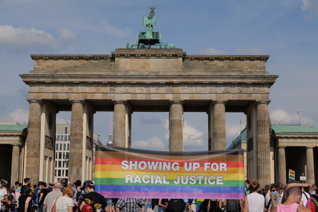 Gruppe von Menschen mit einer "Racial Justice"-Schlagwort vor dem Brandenburger Tor in Berlin, Deutschland, mit den Torpfeilern und der Statue im Hintergrund und Gebäuden und bewölktem Himmel.