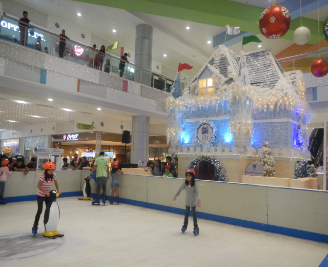 Eine Gruppe von Menschen, die auf einer Eisbahn in einem Einkaufszentrum Schlittschuh laufen, umgeben von einem hausähnlichen Aufbau, der mit Lichtern und festlichen Dekorationen geschmückt ist, mit Menschen in der Nähe und Lichtern an der Decke.