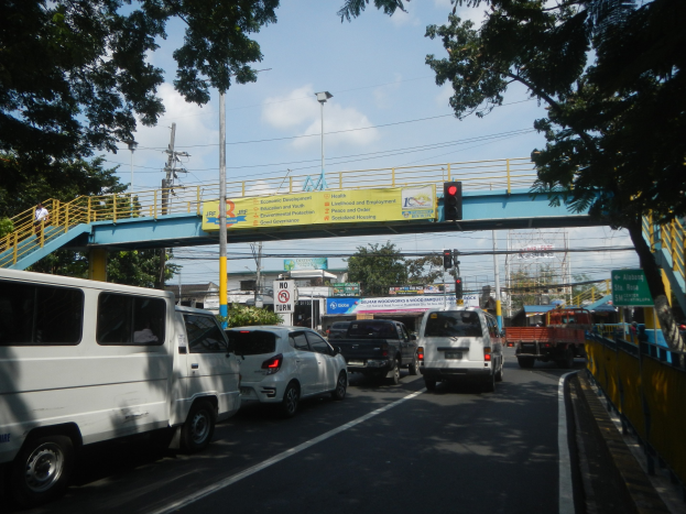 Eine belebte Straße mit Fahrzeugen, eine Brücke mit Geländern und Treppen, Laternen, Verkehrsampeln, Schilder, Bäume, Gebäude und ein bewölkter Himmel im Hintergrund.