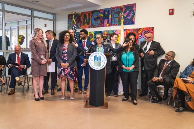 Gruppe von Menschen in einem Raum mit einem Podium, Mikrofon und einer Tafel mit der Aufschrift "Sancho Stadtrat tritt dem Stadtrat bei", zusammen mit Stühlen, stehenden Zuschauern, Fahnen, einem Feuerlöscher und Deckenleuchten.