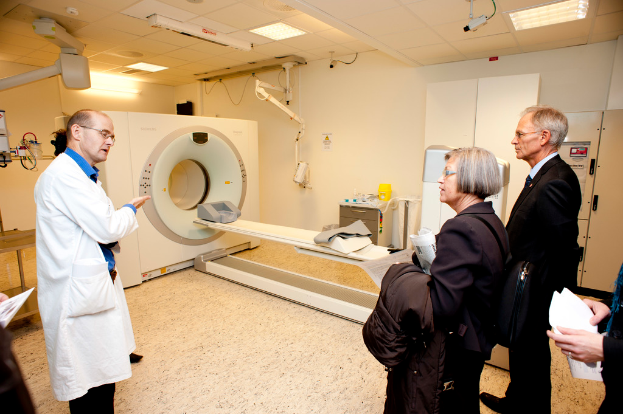 Ein Arzt in weißer Uniform erklärt eine CT-Scan-Maschine zwei Personen in schwarzen Mänteln, die aufmerksam zuhören.