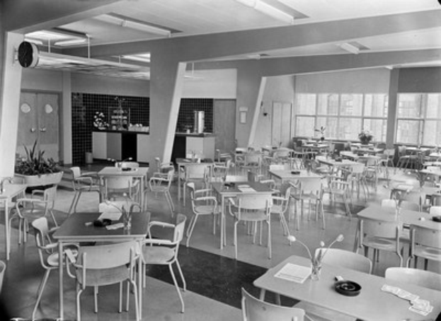 Schwarz-weißes Foto einer Cafeteria mit Tischen und Stühlen, Tellern, Besteck, Gläsern und Blumenvasen auf den Tischen, Türen und Fenstern an der Wand im Hintergrund und Deckenleuchten.