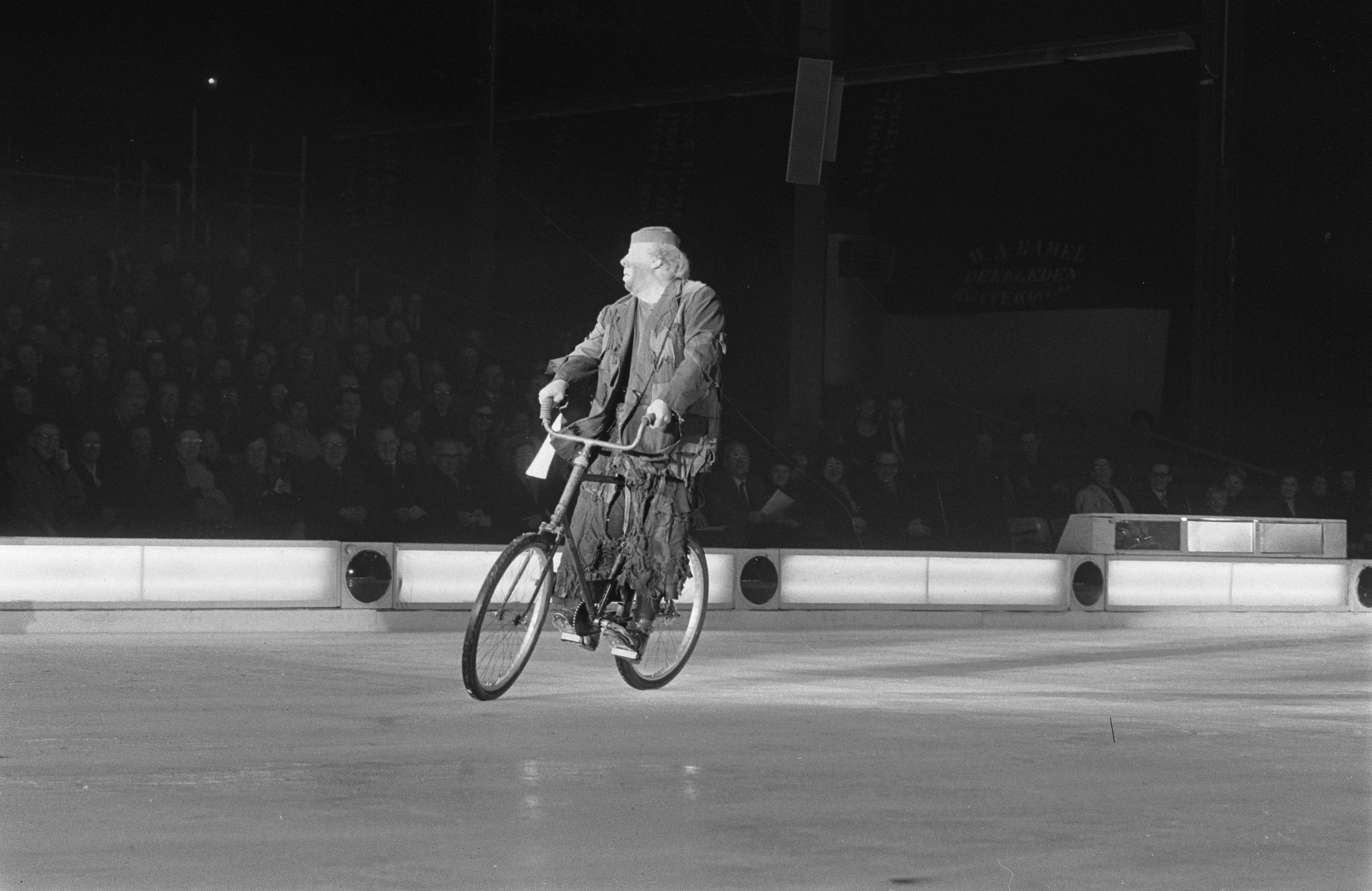 Schwarzes und weißes Foto eines Mannes, der auf einer Eisbahn Fahrrad fährt, mit einer Gruppe von Menschen im Hintergrund und einer Wand dahinter.
