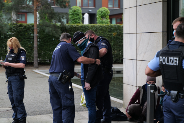 Eine Gruppe von Polizeibeamten vor einem Gebäude mit einer Frau, die ein Buch und einen Stift hält, während Menschen in der Nähe sitzen und stehen, mit Bäumen und Gebäuden im Hintergrund.