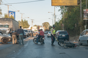 Eine Gruppe von Menschen umringt ein verunglücktes Motorrad am Straßenrand mit mehreren Fahrzeugen, darunter ein Lastwagen, und einer Hintergrundlandschaft aus Bäumen, Pfählen, Laternen und Schildern unter dem Himmel.