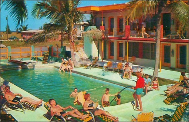 Ein Schwimmbad mit Menschen auf Stühlen, Gebäuden, Bäumen und Autos im Hintergrund.