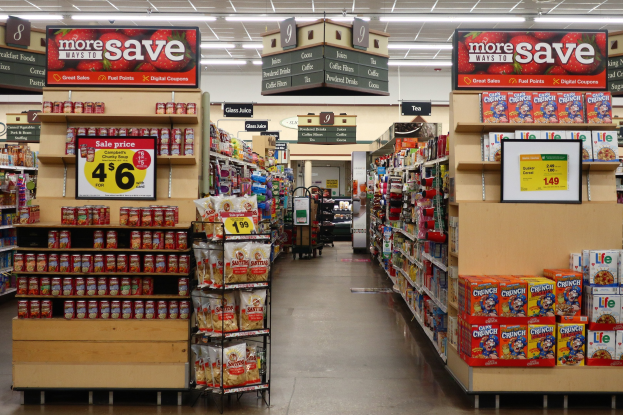 Ein Kiosk-Innenraum mit Regalen und Boden voller verschiedener Lebensmittel, Deckenbeleuchtung und Informationsschildern, mit einer Person im Hintergrund.