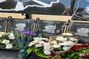 Ein Strauß aus farbenfrohen Blumen ruht auf einem Gehweg neben einem Schild, mit mehreren Gedenktafeln im Hintergrund.