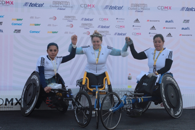 Drei Frauen im Rollstuhl auf einem Podium, die Hände erhoben, in weißen und schwarzen Kleidern und Medaillen, vor einem weißen Banner.