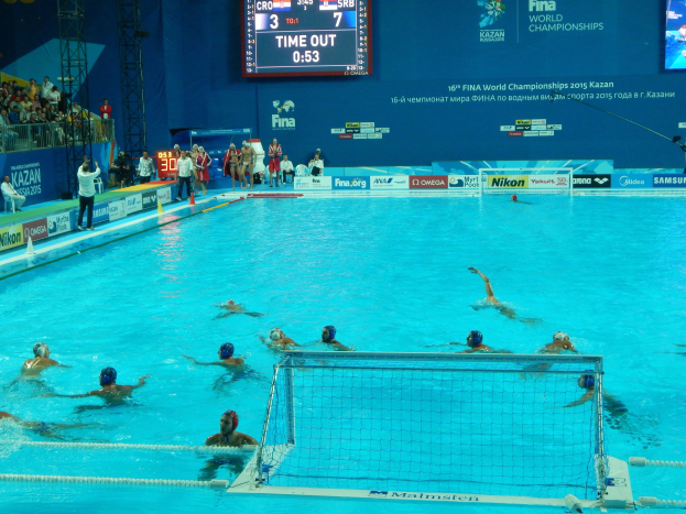 Gruppe von Menschen, die Wasserball in einem Pool während der FINA-Weltmeisterschaften 2015 in Kazan spielen, mit Zuschauern und Eventwerbung im Hintergrund.