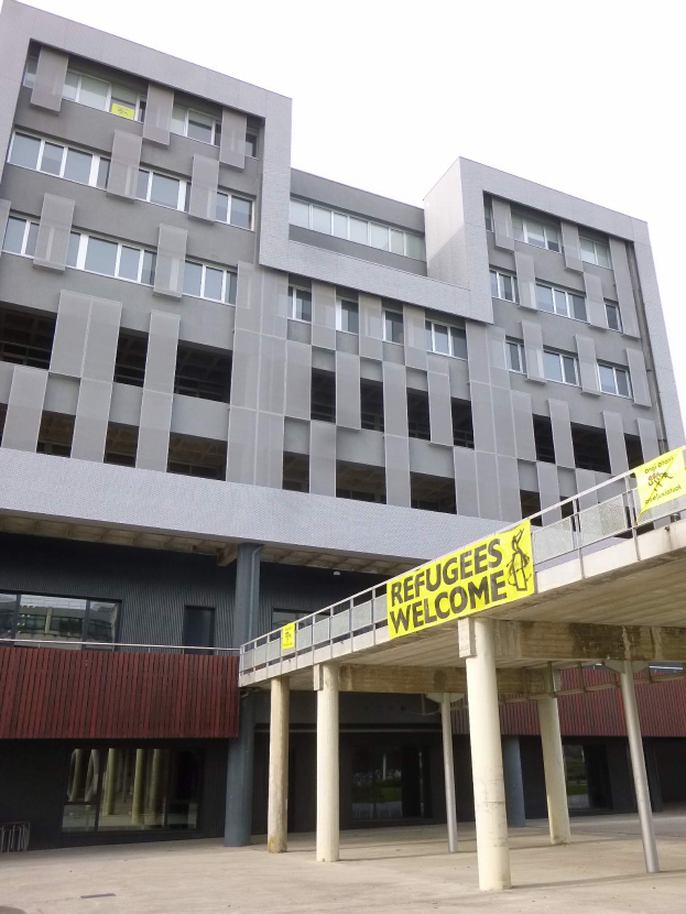 Ein großes Gebäude mit einem Schild "Flüchtlinge Willkommen" davor, umgeben von Säulen und einem Geländer, mit dem Himmel im Hintergrund.