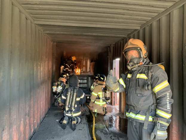 Feuerwehrleute in Schutzausrütstüten innerhalb eines Containers während eines Feuerlöschbetriebs, mit einem haltenden Rohr und einem Feuerlöscher im Hintergrund.