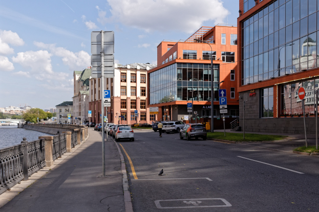 Stadtstra├če mit parkenden Autos, Geb├Ąuden, Schildern, Gras, ├ťberwasserbrücke, B├Ąumen und bew├Âlktem Himmel.