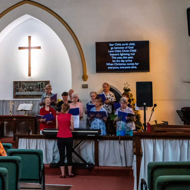 Eine Gruppe von Menschen steht vor einer Kirche, einige halten Bücher, Stühle links, Kreuzsymbol und Foto auf der Wand, ein Schild mit Text, Lautsprecher und ein Tisch mit einer Decke, scheinbar singend als Chor.
