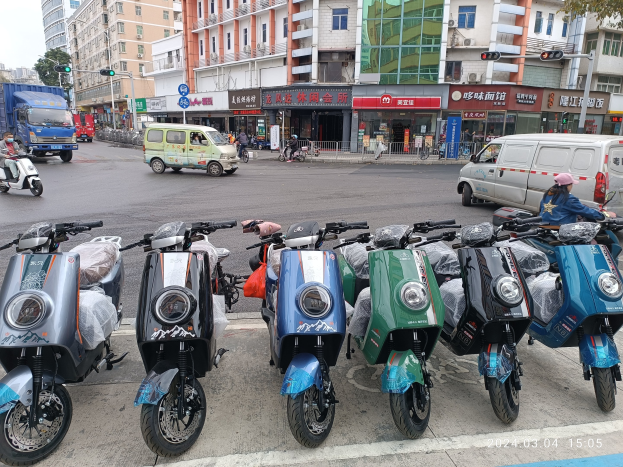 Eine Reihe von elektrischen Rollern, die auf der Straße geparkt sind, mit einer Person in einer Mütze davor, vor einem Hintergrund von Gebäuden, Verkehrsampeln, Bäumen und einem klaren blauen Himmel.