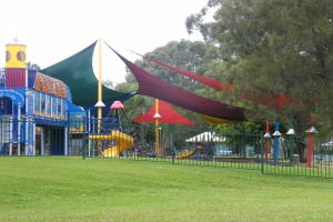 Ein Park mit einem Spielplatz, der von einem Metallzaun umgeben ist und Kinder auf einer Rutsche, Stangen und einem Zelt spielt, vor grünem Gras, Bäumen und einem klaren blauen Himmel.
