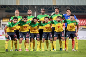 Eine Gruppe von Männern steht auf einem Fußballfeld, feiert einen Sieg und ist von Werbetafeln, Stühlen, Geländern, Pfosten, Bäumen und einer Hütte im Hintergrund umgeben.