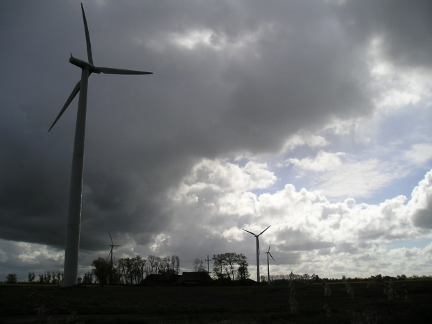Windmühlen mit Bäumen mit Zweigen und Blättern im Vordergrund, Wolken am Himmel und ein Haus im Hintergrund.