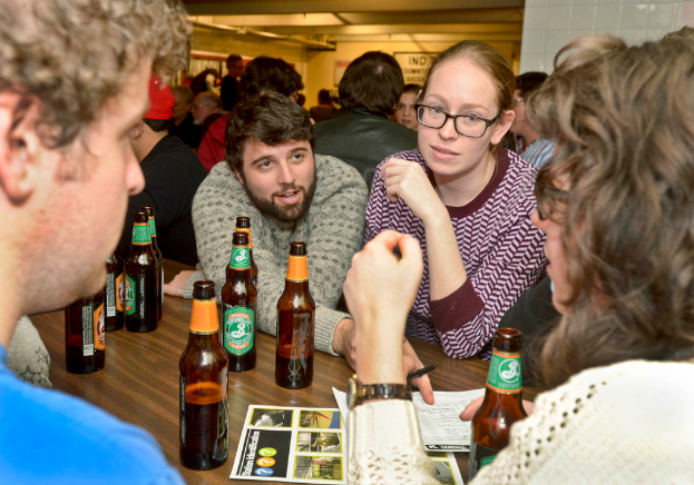 Mehrere Personen auf beiden Seiten eines Tisches mit Bierflaschen in einem Raum, der wie eine Bar aussieht.