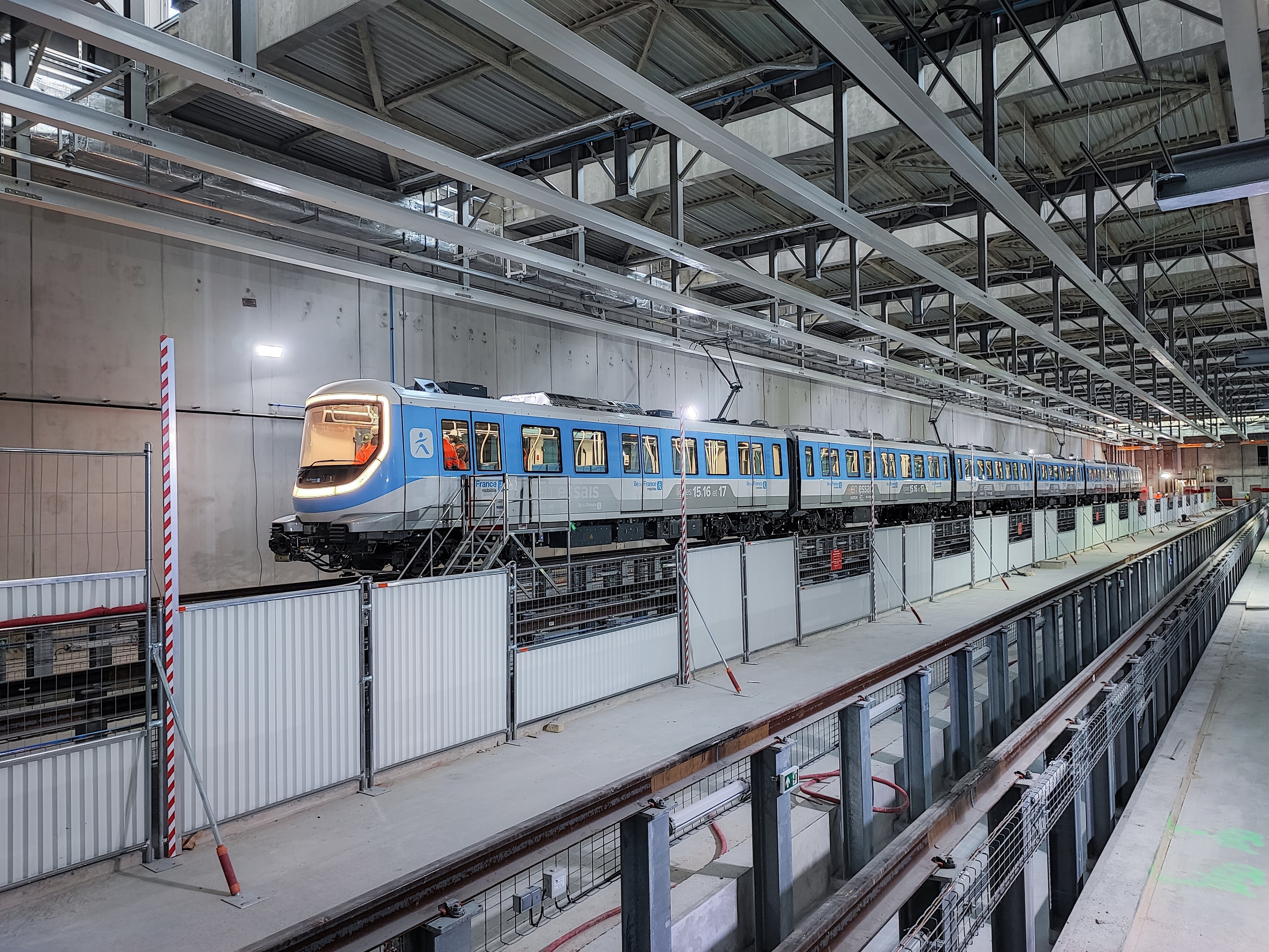 Ein blauer und weißer Zug, der durch eine große Indoor-Station fährt, mit einer Plattform auf der rechten Seite und einer Wand im Hintergrund, mit an der Decke montierten Stangen und Lampen.