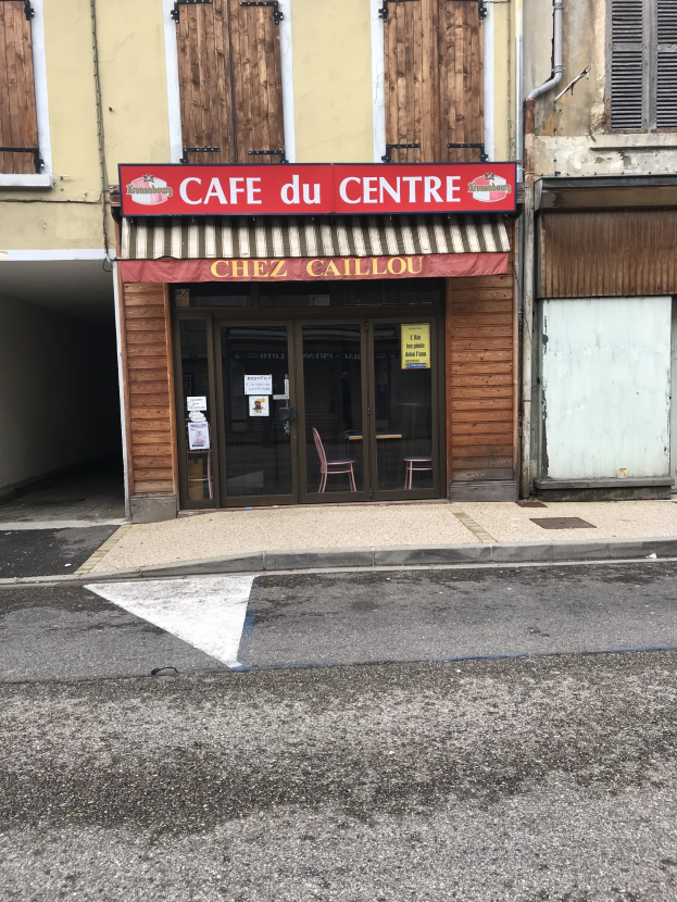 Außenansicht eines Gebäudes mit einer 'Café du Centre'-Tafel, Fenstern, einer Tür und einer Straße davor mit einer Texttafel.